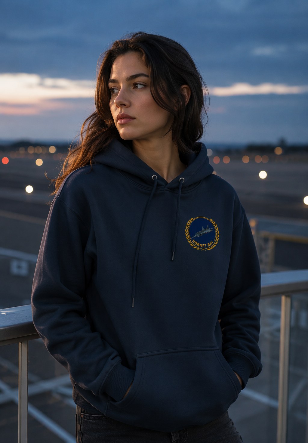 Model at a dusk runway overlook wearing the navy Squadron Laurel Hoodie with the laurel emblem on the left chest